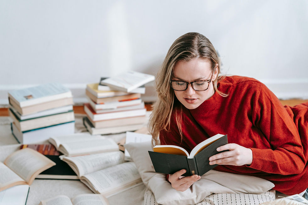 Woman in red sweater, glasses, reading a book on the floor with scattered books around her. Relaxed, cozy setting with white walls.