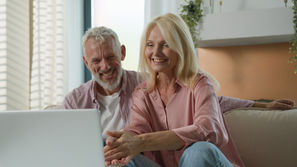 Smiling couple sitting on a couch, using a laptop. Bright room, plants in background. Both wearing pink shirts, conveying a joyful mood.