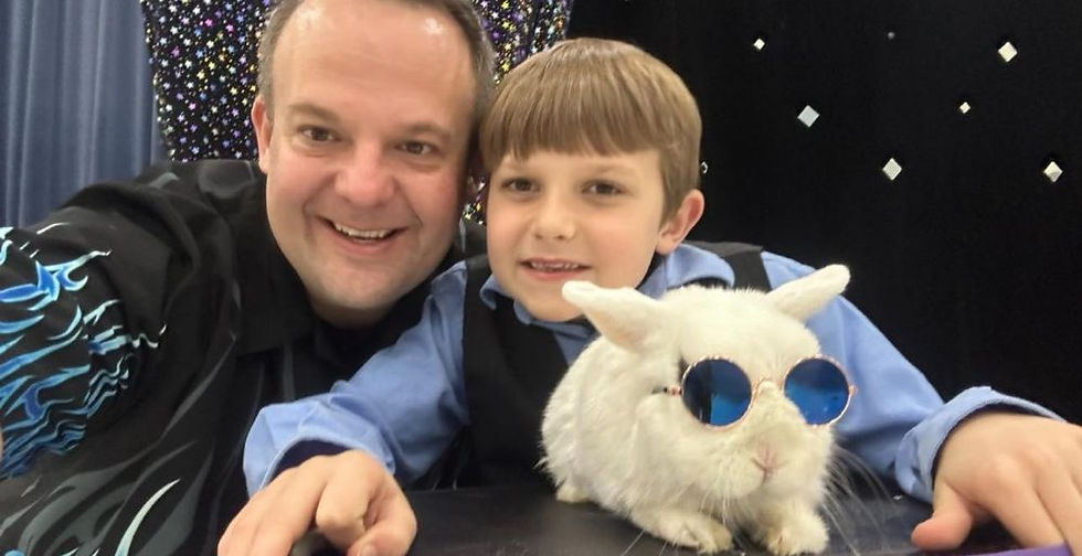 Man and boy smiling with a white rabbit wearing blue sunglasses. They are indoors against a sparkling black background, creating a joyful scene.