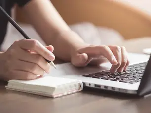 Hands resting on a desktop computer and a pen being held in the right hand