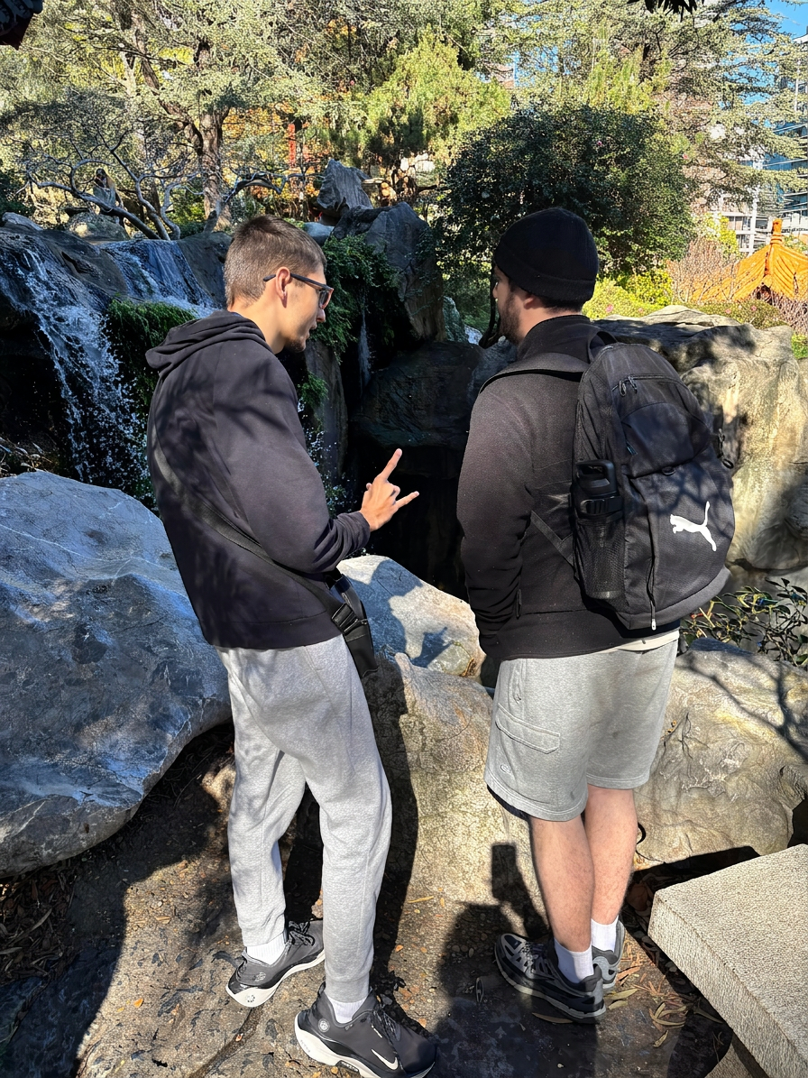Outdoor photography of two young men conversing by a waterfall in a park setting. Surrounded by rocks and greenery, the scene suggests a relaxed, sunny atmosphere.