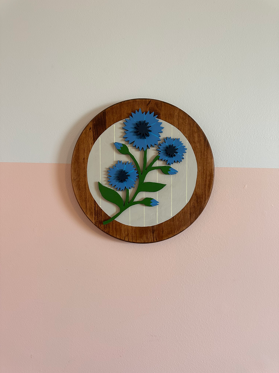 Thumbnail: Circular wooden plaque with blue flowers against a white and pink wall.
