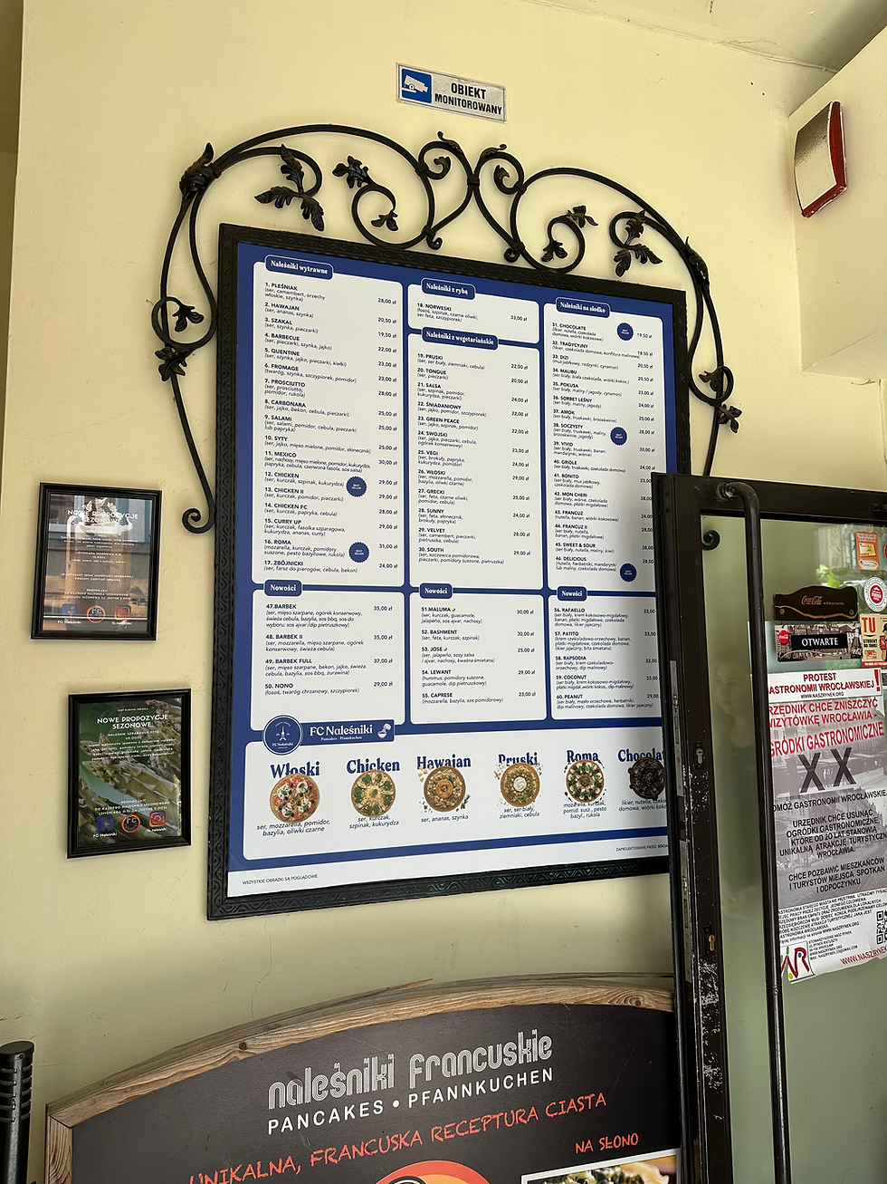 Niebiesko-białe menu naleśników na ścianie restauracji, ozdobiona metalową ramą; obok znak Obiekt monitorowany i plakaty.