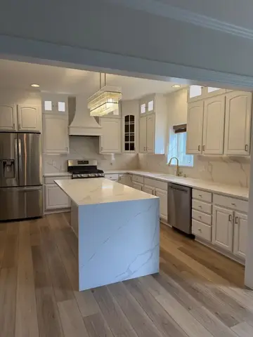 Kitchen with cabinets painted white, lit glass-front toppers, quartz waterfall island, full-height quartz backsplash, crystal chandelier and plank floor, Macedonia, Ohio
