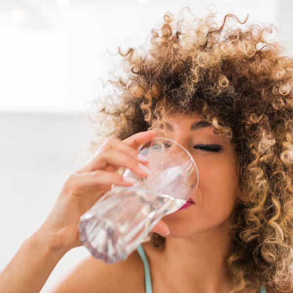 Woman with curly hair drinking water from a glass. Bright setting, relaxed mood, wearing a light blue strap top.