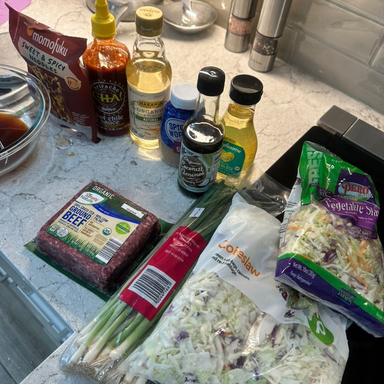 Ingredients on a kitchen counter include coleslaw, green onions, ground beef, sauces, oil, and seasoning. Bright, organized setup.