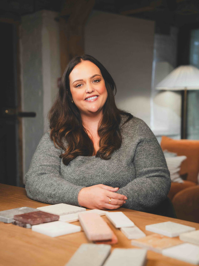 A woman posing for a photo at a desk surrounded by various interior design materials as she leads a design meeting with a client.