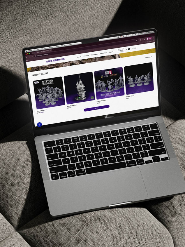 A laptop is resting on a sofa showing Chaos Realm Prints website featuring 3D printed miniatures. 