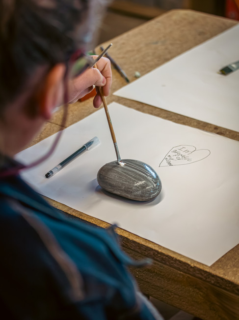 An individual painting on a grey, oval stone at a workshop table, with a pen and a sketch visible in the background, engaging in a creative craft activity.