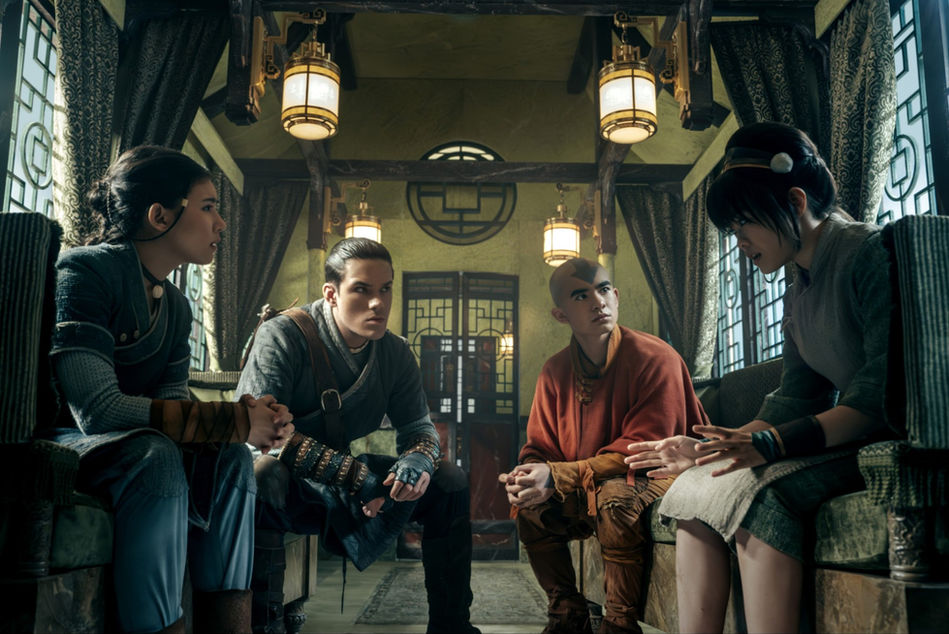 A medium wide image of 4 young teenagers, 2 females and 2 males, sitting in a stylish passenger train.  One of the females is talking as the other 3 listen intently and in secrecy.