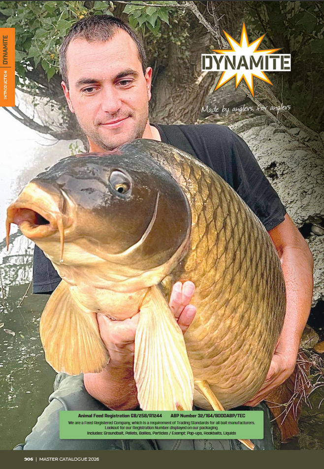 Pêcheur tenant une grande carpe commune dorée sortie de l’eau, photographié au bord d’une berge boisée. En arrière-plan, feuillage et reflet de l’eau. En haut à droite figure le logo Dynamite Baits accompagné du slogan « Made by anglers, for anglers ».