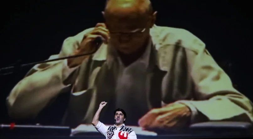 Imagine João Suassuna, historiador e produtor cultural, em um palco iluminado, apresentando uma aula-espetáculo sobre seu avô, Ariano Suassuna. Ele está envolvido em uma narrativa cativante, compartilhando histórias e reflexões sobre a vida e obra do renomado dramaturgo. O público está atento, absorvendo cada palavra e gesto, enquanto João utiliza recursos visuais e musicais para enriquecer a apresentação. A atmosfera é de admiração e respeito pelo legado cultural de Ariano Suassuna.