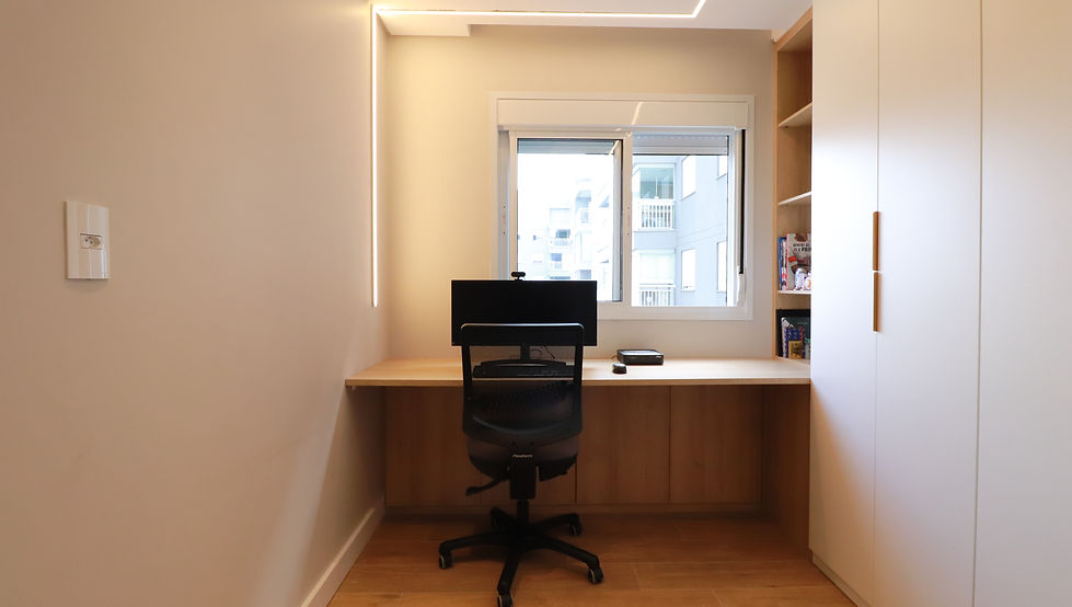 Escritório residencial do Apartamento IR01, no Jardim Marajoara, São Paulo, projetado pela GUZLIVE. O ambiente apresenta bancada de trabalho em madeira sob a janela, marcenaria planejada branca com nichos e armários, cadeira ergonômica preta, piso amadeirado e iluminação linear em LED, criando um home office contemporâneo e funcional, planejado para futura conversão em quarto de bebê.