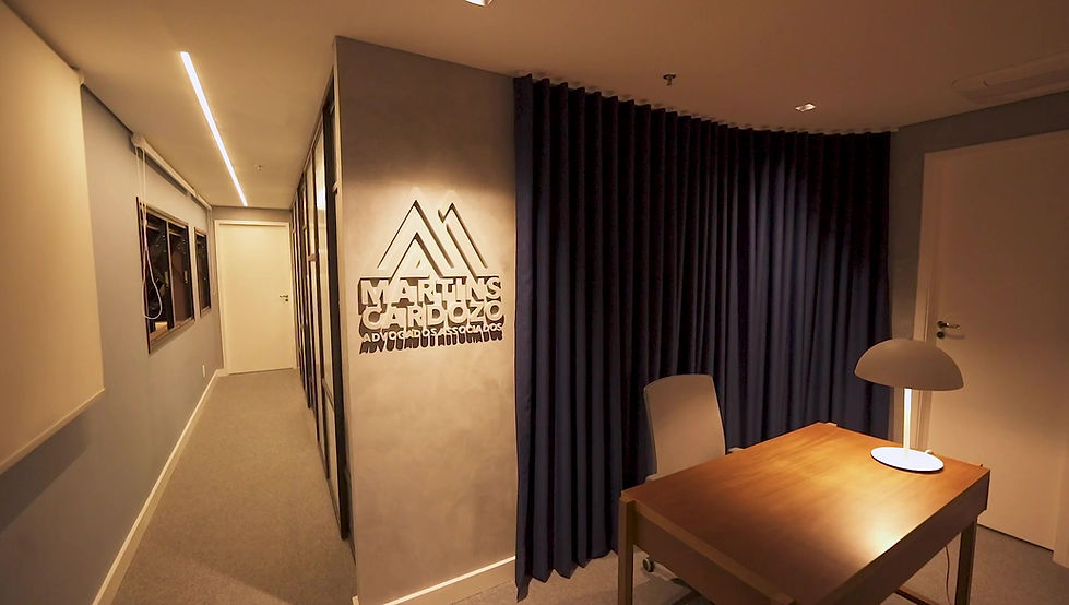 Recepção do escritório Martins Cardozo Advogados, em São Paulo, projetada pela GUZLIVE, com mesa de madeira modernista, luminária de design, cortinas azuis, logotipo na parede em cimento queimado e corredor com divisórias de vidro, combinando arquitetura corporativa contemporânea, privacidade e iluminação acolhedora.