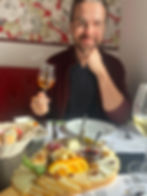 Man smiling, holding a glass of wine at a table with a cheese platter. Floral wallpaper, red seating, cozy atmosphere.