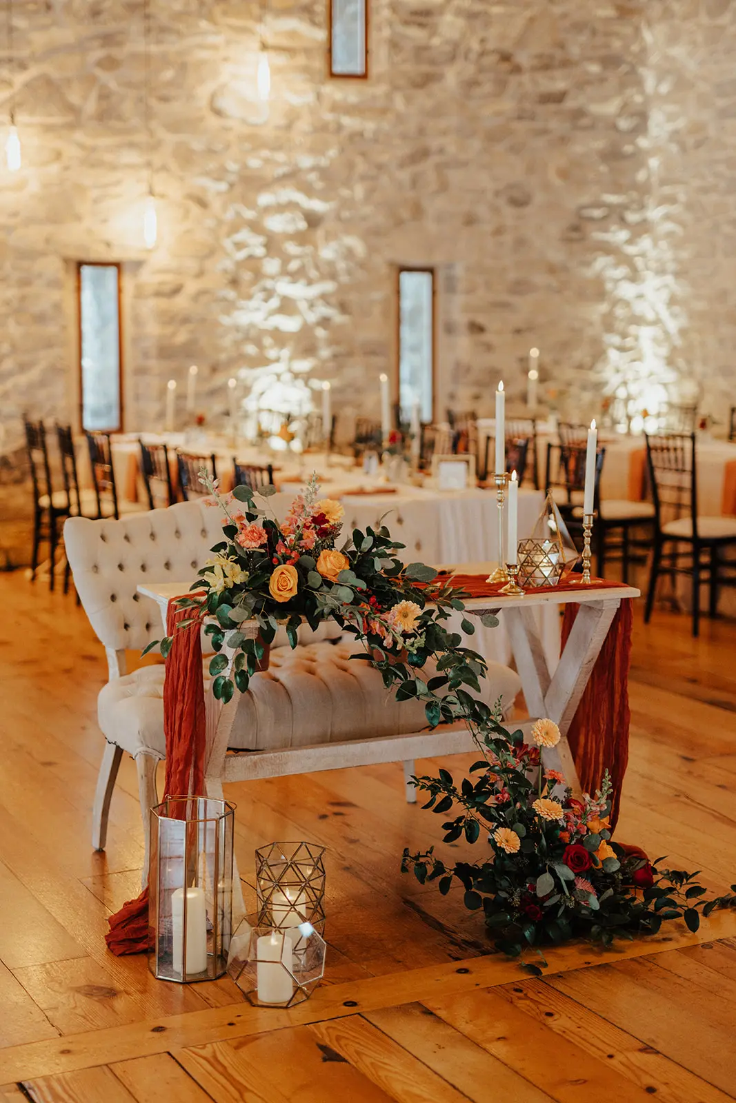 Elegant wedding reception table with warm uplighting, red and orange floral arrangements, and flowing drapes in a historic venue with stone walls.