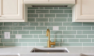 A modern Kitchen backsplash with light green tiles, off-white countertop, golden faucet an