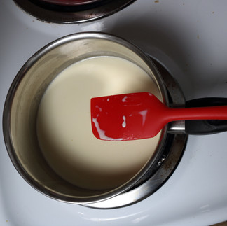 Small pan of heavy cream sits on a stove burner, heating up.  There is a red spatula resting on the handle of the pan, ready to give the cream a stir so it doesn't burn.