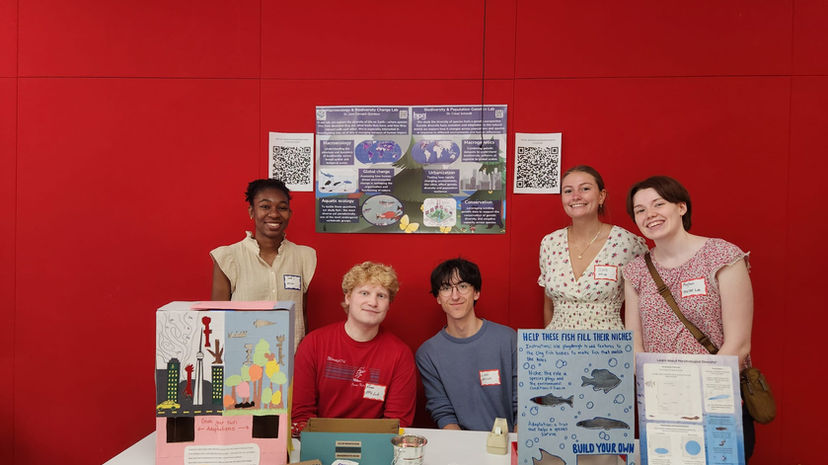 A picture of 5 students at a booth with a poster in the background. On the table are city and ocean dioramas for people to create animals with playdoh to fill.