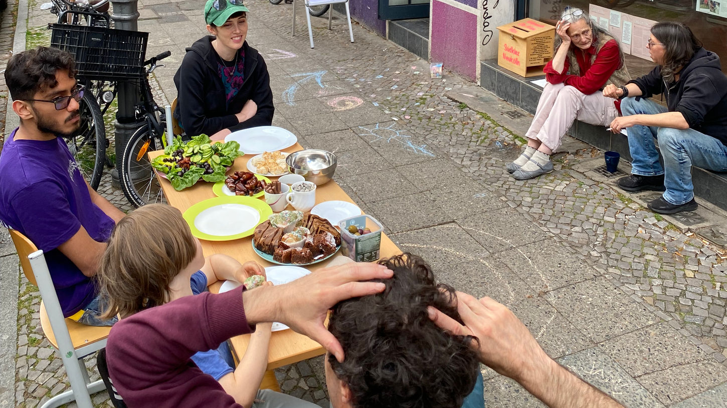 Gruppe sitzt an einem Tisch mit Essen vor einer violetten Fassade Kiezbegegnung.