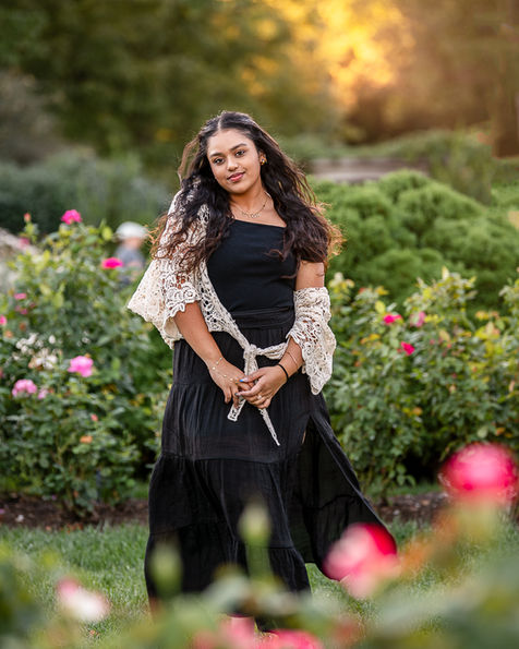 Senior girl in a black dress at golden hour in a rose garden.