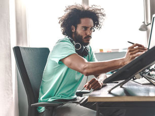 Man in teal shirt and headphones draws on a pen display at a desk, focused in a bright office with a computer monitor.