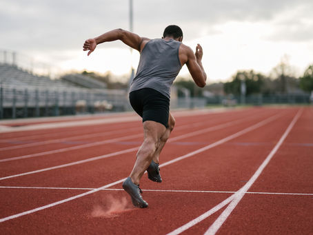 Sprinter at maximal velocity photographed from behind and slightly to the side at track level, with one foot loaded against the surface and the opposite leg driving through.