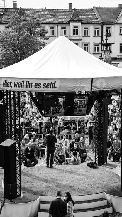 Menschenmenge hört Sprecher unter Zelt zu, Banner: Hier, weil Ihr es seid.