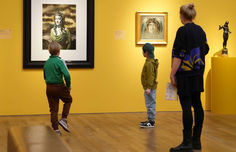 Zwei Grundschulkinder in lockeren Pullis blicken zusammen mit der Mutter auf die Exponate der Kunstausstellung - von hinten fotografiert