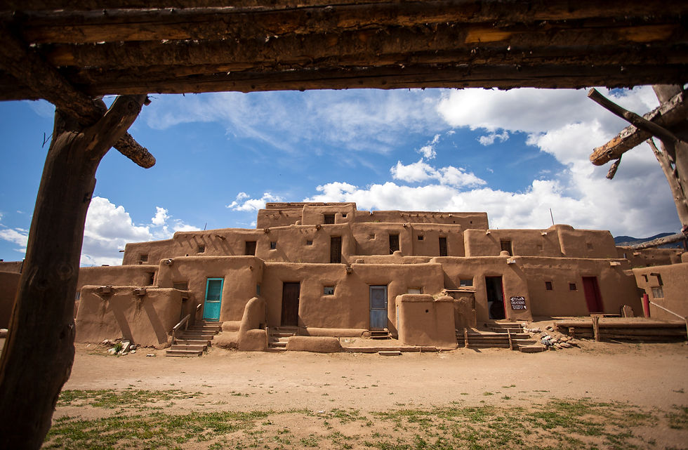 Adobe pueblo buildings under a blue sky, framed by wooden beams; turquoise door and a small sign, quiet desert scene