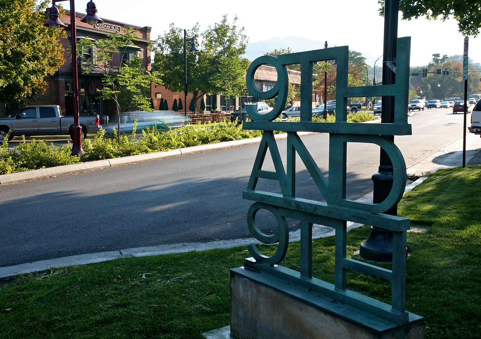 Green metal 9th and 9th sculpture on a sunny street beside the Woodman Bldg, with trees, parked cars, and traffic.