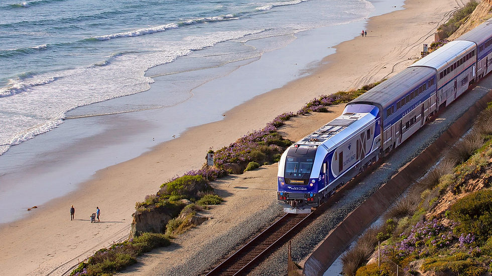 Train travels along a scenic coastline with a sandy beach and ocean waves. People walk nearby; vibrant greenery and flowers line the track.