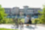Couple sits on a bench by a riverfront, with two parked bikes in front and storefronts behind.