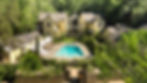 Aerial view of a yellow hillside estate with a turquoise pool, white umbrellas, and lush green trees surrounding the complex.