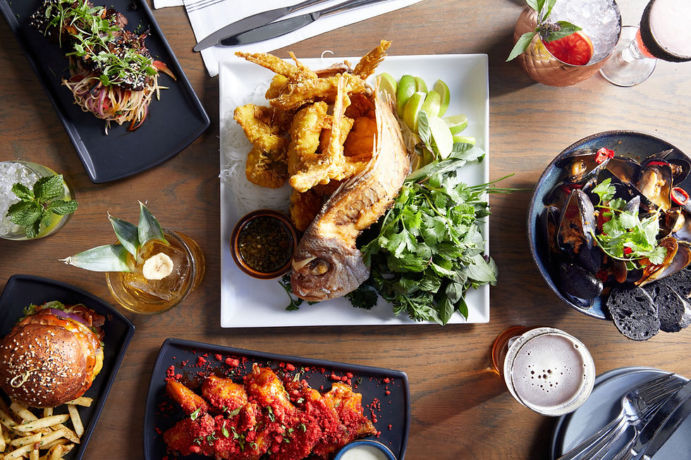 An assortment of dishes on a wooden table, including fried fish with herbs, mussels, a burger with fries, and colorful drinks.