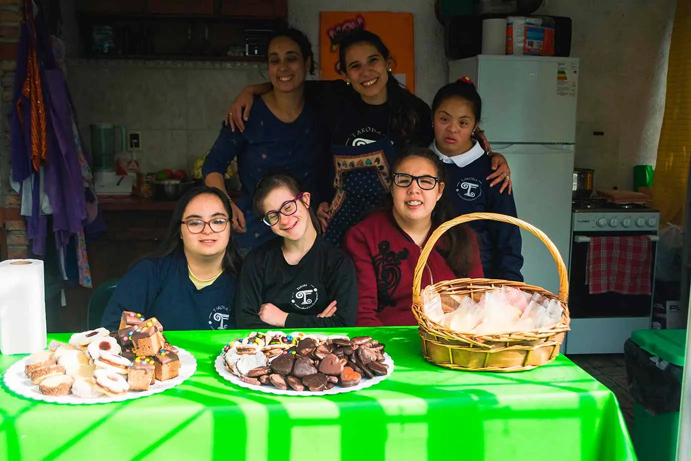 Personas con síndrome de down, autismo y sin discapacidad detrás de una mesa con masitas y tortas a la venta.