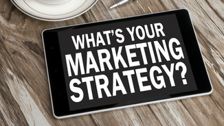 Tablet on wooden table displaying the text "WHAT'S YOUR MARKETING STRATEGY?" alongside a pen and saucer, conveying a strategic mood.