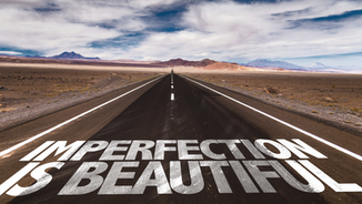 Open road in desert, stretching to horizon under cloudy sky. Bold white text on road reads "IMPERFECTION IS BEAUTIFUL," evoking inspiration.