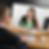 Man in suit video calls smiling woman on monitor at wooden desk. Webcam, mic, and call icons visible. Bright, professional setting.