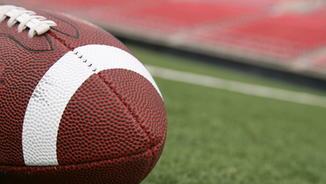 Close-up of a football on green turf, blurred red stadium seats in the background. The scene is calm and focused on the ball.