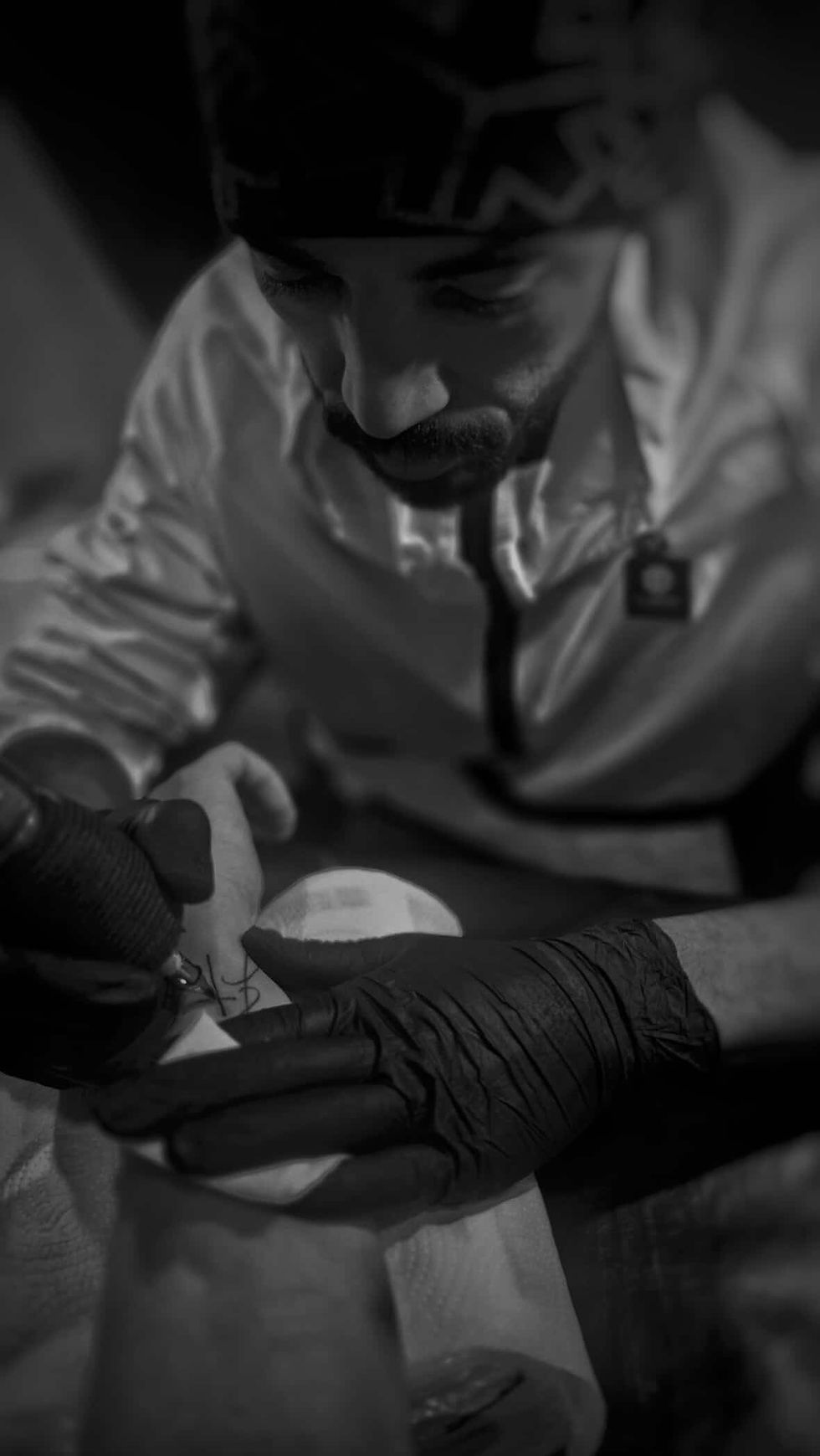 Foto em preto e branco, vertical, de tatuador trabalhando em close nas mãos, com luvas pretas e foco na agulha, fundo desfocado.