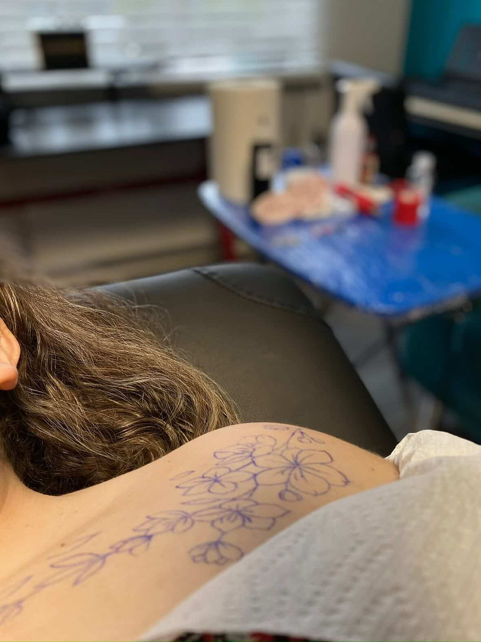 Foto horizontal de cliente deitada na maca com decalque de tatuagem floral em fine line no ombro