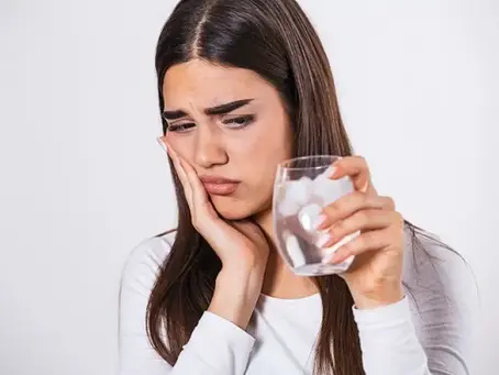 امرأة ممسكة بكوب ماء بارد بيدها، تعبير وجهها يعكس ألمًا نتيجه حساسية في الأسنان. ترتدي قميصًا أبيض، الخلفية بيضاء، الجو متوتر.