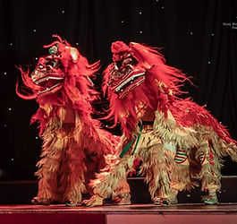 Two red Chinese lion dance costumes with performers inside appearing mid-performance on a theatre stage with dark backdrop and spotlights