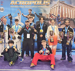 China Spirit UK team posing at the Acropolis International Wushu Open Tournament in Athens, wearing medals and holding several large trophies in front of the event backdrop, with athletes standing and kneeling together on the competition floor.