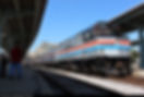 Silver Amtrak train at a station, people on the platform. Bright blue sky, train has red and blue stripes. Calm, sunny day.