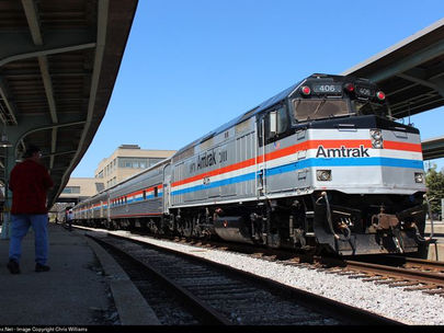 Silver Amtrak train at a station, people on the platform. Bright blue sky, train has red and blue stripes. Calm, sunny day.