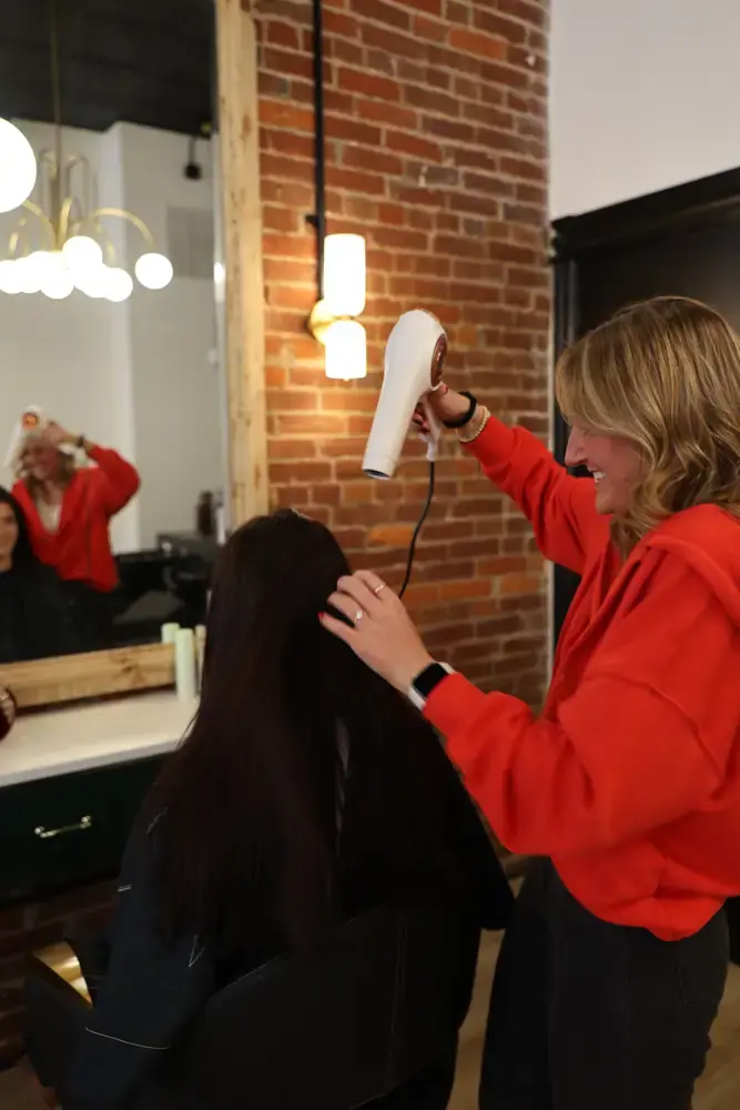 Stylist finishing a blowout on long dark hair beside exposed brick at Aura Hair Studio in Lawrenceville, Pittsburgh