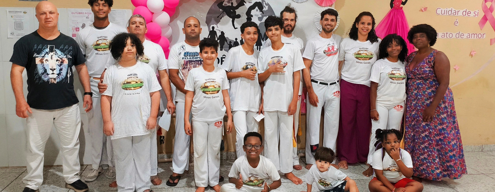 Integrantes da Associação AIDÊ, entre mulheres, crianças e educadores de capoeira, posam diante do mural da campanha Outubro Rosa 2025, com a mensagem ‘Cuidar de si é um ato de amor’. A ação simboliza a união entre cultura, saúde e autocuidado na comunidade de Ipatinga.