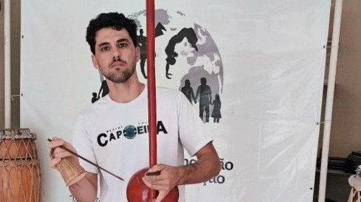 Graduado Miau segura um berimbau diante da identidade da AIDÊ em aula de 5 de setembro de 2026.